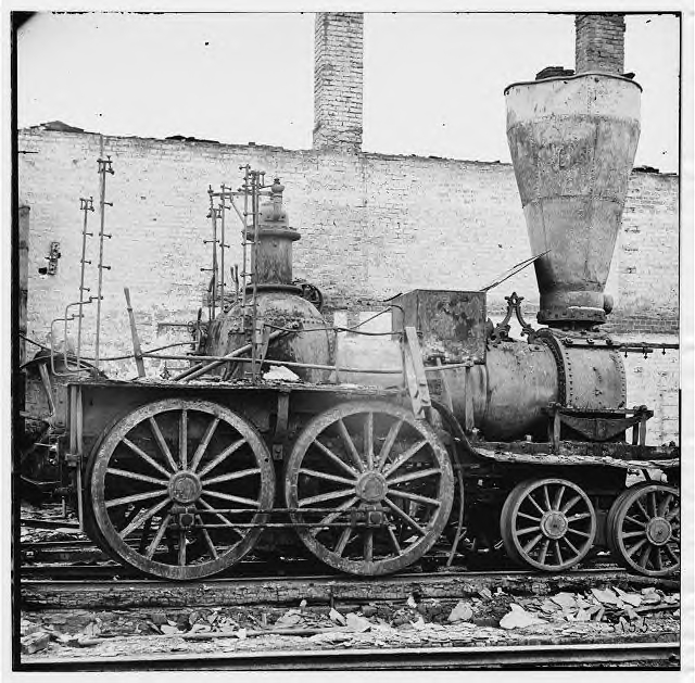 A destroyed locomotive captured at Richmond's fall. Many locomotives this badly burned up were repaired by the Confederate railroads, though with great difficulty in finding the required replacement parts. The fire has removed the woodwork and shows the wheels, frame and steam generation system. To the left is the firebox; on the right is the smoke chamber and stack; between is the horizontal boiler.